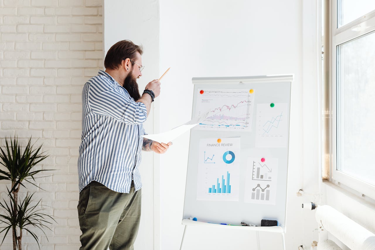 A man with a beard examines financial charts on a whiteboard in an office setting.